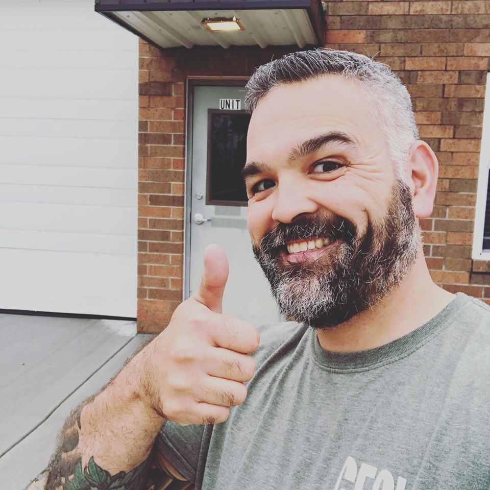 A bearded man smiles and gives a thumbs up while standing outside a brick building with a door labeled Unit 1.
