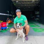 A smiling man in a green UNICORN LOVER shirt and black cap is kneeling on a tiled floor, hugging a black and white dog, inside a large garage with cleaning supplies in the background.