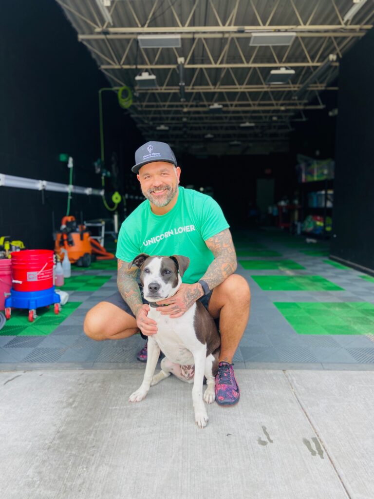A smiling man in a green UNICORN LOVER shirt and black cap is kneeling on a tiled floor, hugging a black and white dog, inside a large garage with cleaning supplies in the background. A smiling man in a green UNICORN LOVER shirt and black cap is kneeling on a tiled floor, hugging a black and white dog, inside a large garage with cleaning supplies in the background.