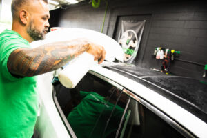 A man with tattoos wearing a green shirt sprays cleaning solution on the roof of a white car inside a detailing shop. Bottles and supplies are visible on the black wall in the background.