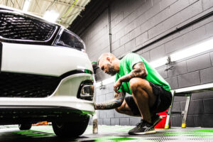 A man in a green shirt crouches beside a white car, carefully cleaning or detailing the front bumper inside a brightly lit garage with black walls.