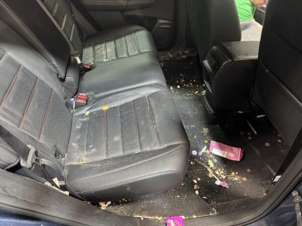 The back seat and floor of a car are messy, with scattered trash, food crumbs, candy wrappers, and coins. The seats are black leather, and a person in a green shirt is partly visible outside the open door.