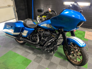 A shiny blue touring motorcycle with black and chrome accents sits on a green and gray checkered floor in a garage with black walls and bright overhead lighting.