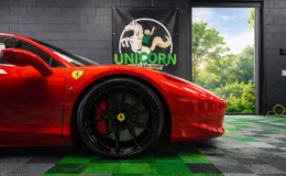 A red Ferrari with black wheels is parked indoors on a green and gray checkered floor. Behind it, there is a Unicorn Details McHenry banner and an open door revealing green trees and blue sky outside—showcasing the flawless finish achieved with ceramic coating in McHenry County.