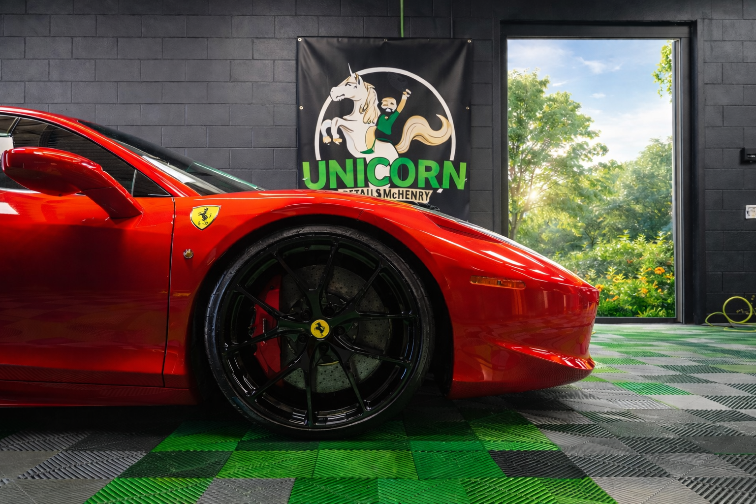 A red Ferrari with black wheels is parked indoors on a green and gray checkered floor. Behind it, there is a Unicorn Details McHenry banner and an open door revealing green trees and blue sky outside—showcasing the flawless finish achieved with ceramic coating in McHenry County.