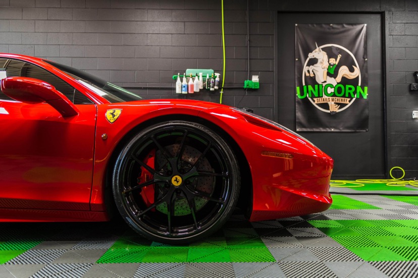 Close-up of a red Ferrari sports car parked indoors, showing the front wheel and part of the hood. In the background, theres a black wall, cleaning supplies, and a Unicorn detailing services banner. Close-up of a red Ferrari sports car parked indoors, showing the front wheel and part of the hood. In the background, theres a black wall, cleaning supplies, and a Unicorn detailing services banner.