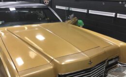 A shiny gold classic car with a black roof is parked indoors. A person in a green shirt is partially hidden behind the car, near the front right side. The wall behind is black with shelves and a logo.
