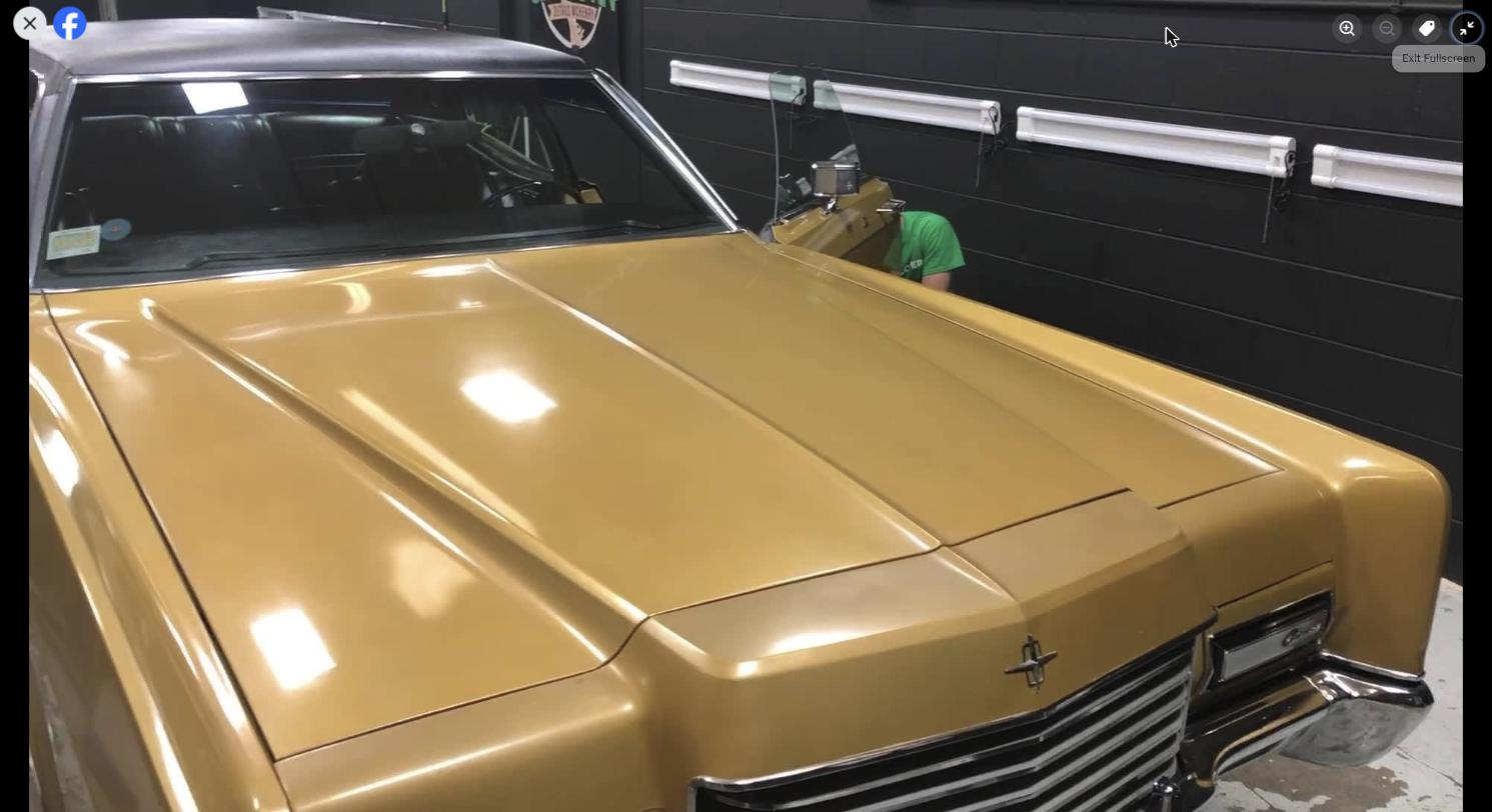 A shiny gold classic car with a black roof is parked indoors. A person in a green shirt is partially hidden behind the car, near the front right side. The wall behind is black with shelves and a logo.
