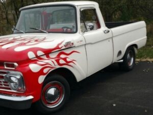 A vintage white pickup truck with red flame decals painted on the hood and sides, parked on a blacktop driveway with green trees and grass in the background.