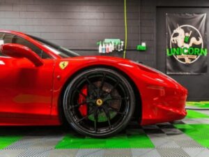 A shiny red Ferrari with black wheels is parked indoors on a green-and-gray checkered floor. In the background, a black wall features car care supplies and a “Unicorn Detailing Services” banner.