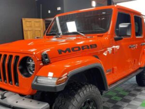 An orange Jeep Wrangler Moab edition is parked indoors on a checkered floor, with the word “MOAB” displayed on the side of the hood. The vehicles front and side are visible.