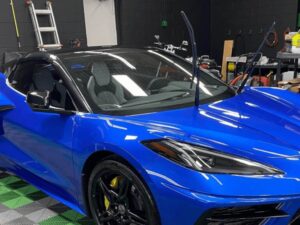 A bright blue sports car with yellow brake calipers is parked indoors. Its windshield wipers are raised, and various tools and equipment are visible in the background, along with a ladder leaning against a black wall.