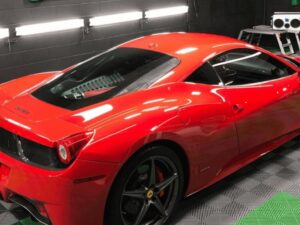 A bright red Ferrari sports car parked indoors on a patterned garage floor, with bright lights mounted on the black wall in the background.