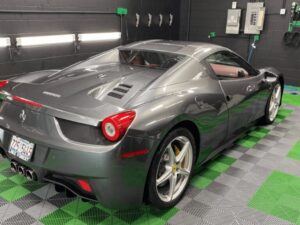 A gray Ferrari sports car is parked indoors on a green and gray checkered floor, with wall-mounted lights, cleaning tools, and electrical panels visible in the background.